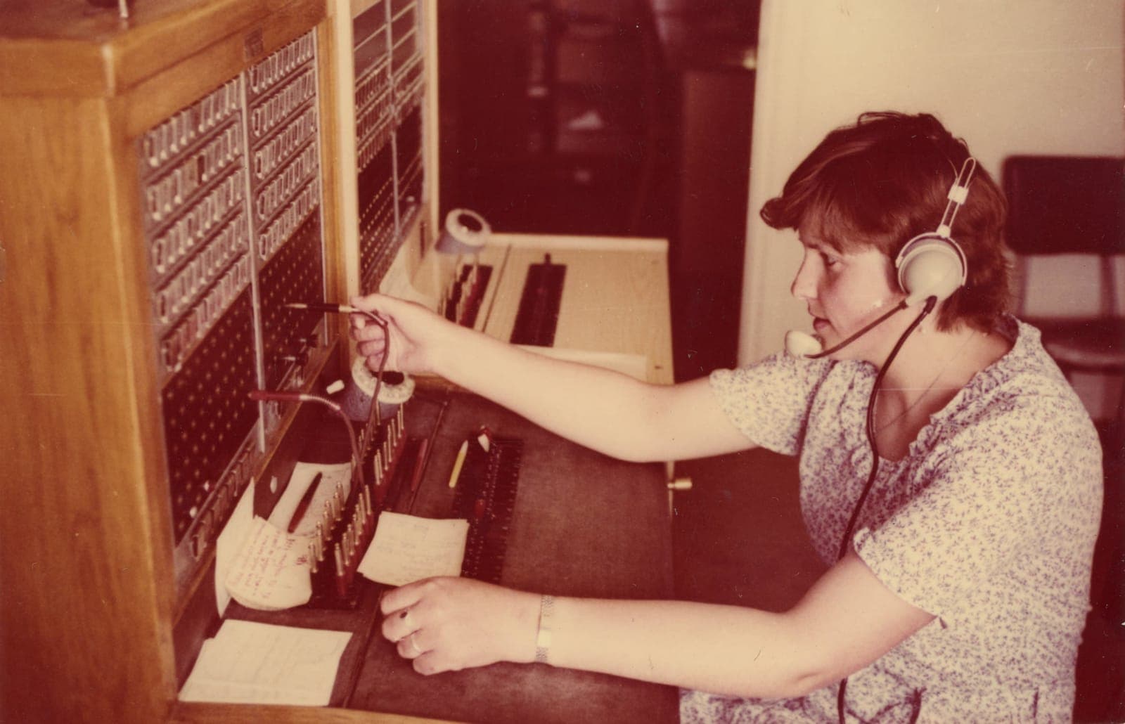 Kobieta w zestawie słuchawkowym obsługuje zabytkową centralę telefoniczną, łącząc połączenia za pomocą kabli i wtyczek, jednocześnie odwołując się do odręcznych notatek na biurku.