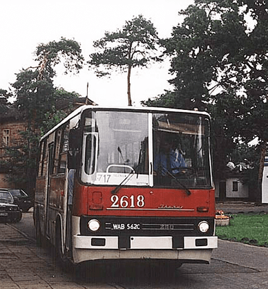 Fotografia kolorowa. Pętla autobusowa. W centralnym miejscu stoi miejski autobus pomalowany na czerwony i biały kolor. Po lewej stronie na chodniku kilka osób z rowerami, po prawej niski budynek, trawa i drzewa