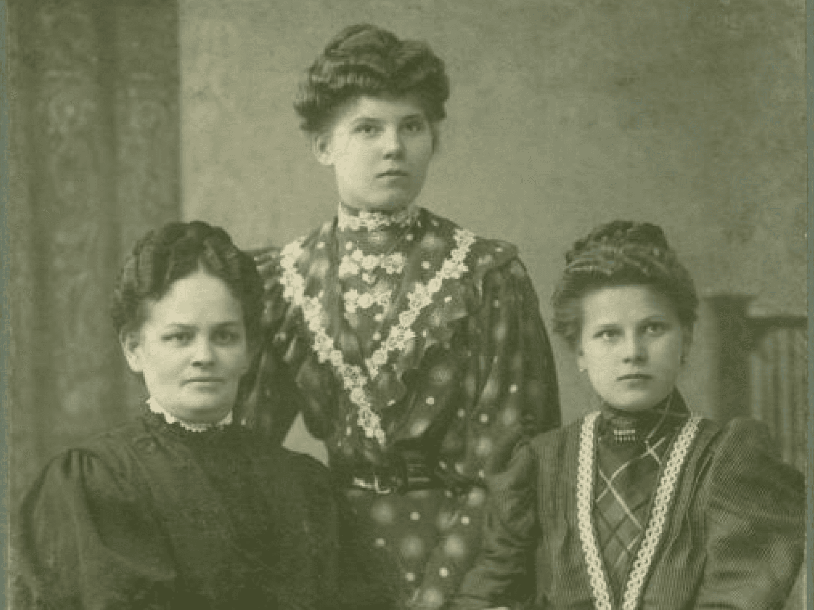 Portret trzech kobiet z początku XX wieku, ubranych w eleganckie długie suknie. Wszystkie kobiety mają upięte włosy. Dwie siedzą przy stole, jedna stoi pomiędzy nimi. Na stole leży obrus i stare pisma. Zdjęcie wykonane w atelier fotograficznym.  Link do strony z artykułem zainspirowanym Zbiorami Społecznymi