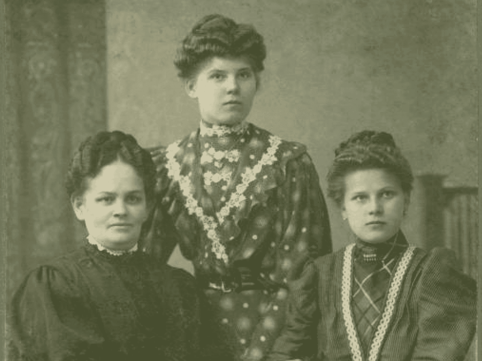 Portret trzech kobiet z początku XX wieku, ubranych w eleganckie długie suknie. Wszystkie kobiety mają upięte włosy. Dwie siedzą przy stole, jedna stoi pomiędzy nimi. Na stole leży obrus i stare pisma. Zdjęcie wykonane w atelier fotograficznym.  Link do strony z artykułem zainspirowanym Zbiorami Społecznymi