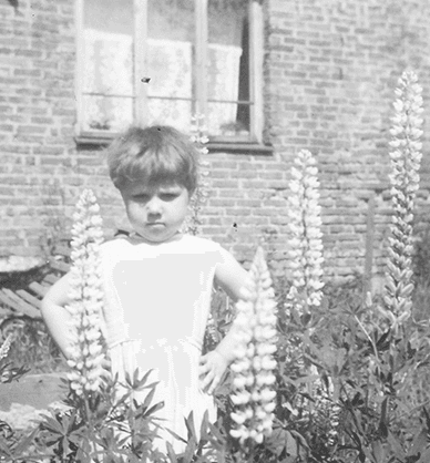 Fotografia czarno-biała. Kilkuletnia dziewczynka stoi w ogrodzie przed domem. Ma nadąsany wyraz twarzy i podpiera się pod boki. Dom z murowanej cegły z dużymi oknami. W jednym oknie wisi biała firanka. W ogrodzie kwitnie bujna trawa i rosną kwiaty łubinu