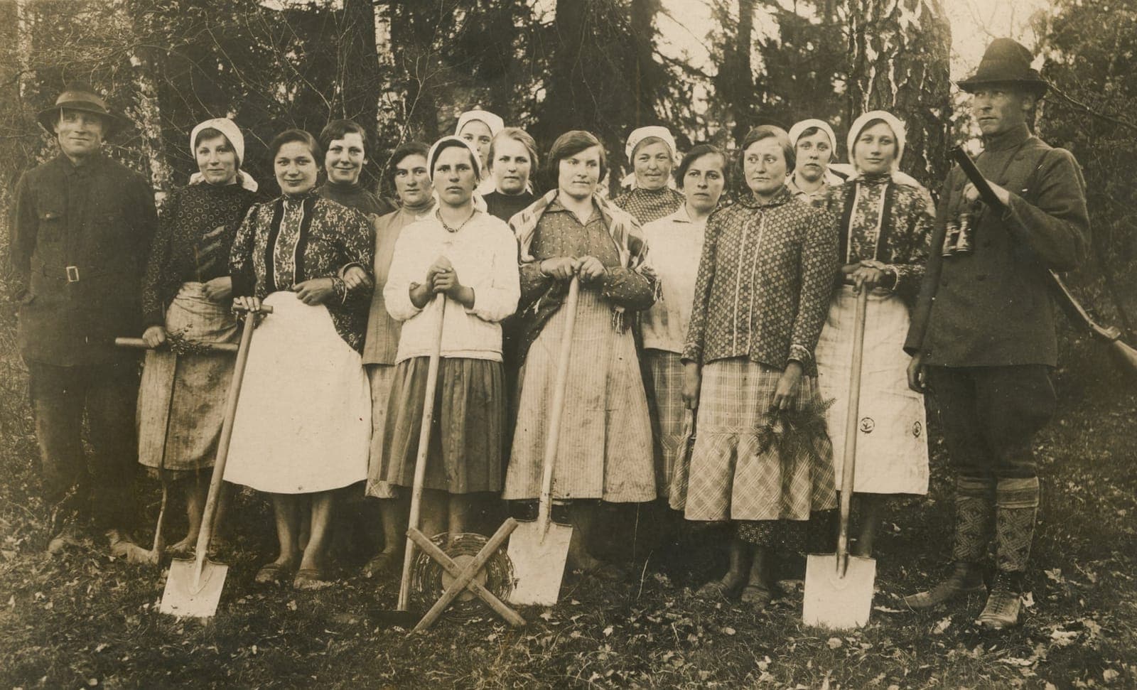 Grupa kobiet trzymających łopaty stoi na świeżym powietrzu między dwoma mężczyznami w mundurach, a w tle widać drzewa.