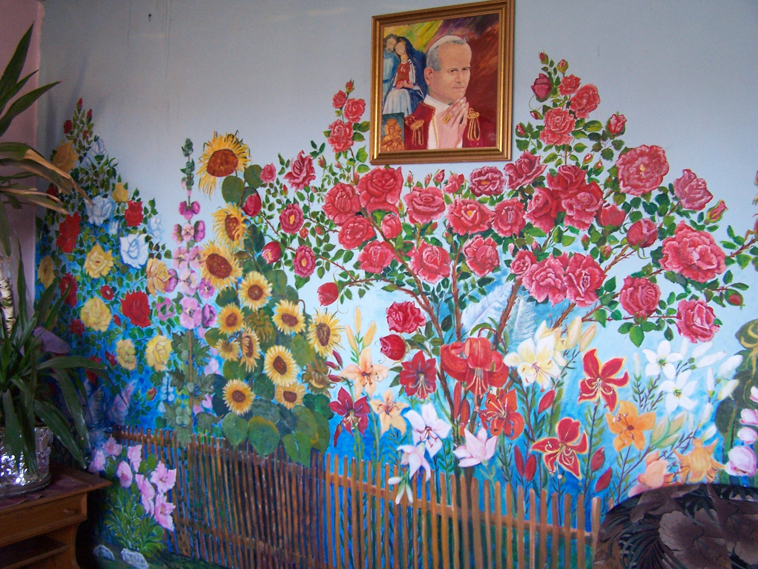 Kolorowy kwiatowy mural ze słonecznikami, różami i liliami pokrywa ścianę. Nad muralem wisi oprawiony portret osoby w czerwieni. Po lewej stronie znajduje się doniczkowa roślina.