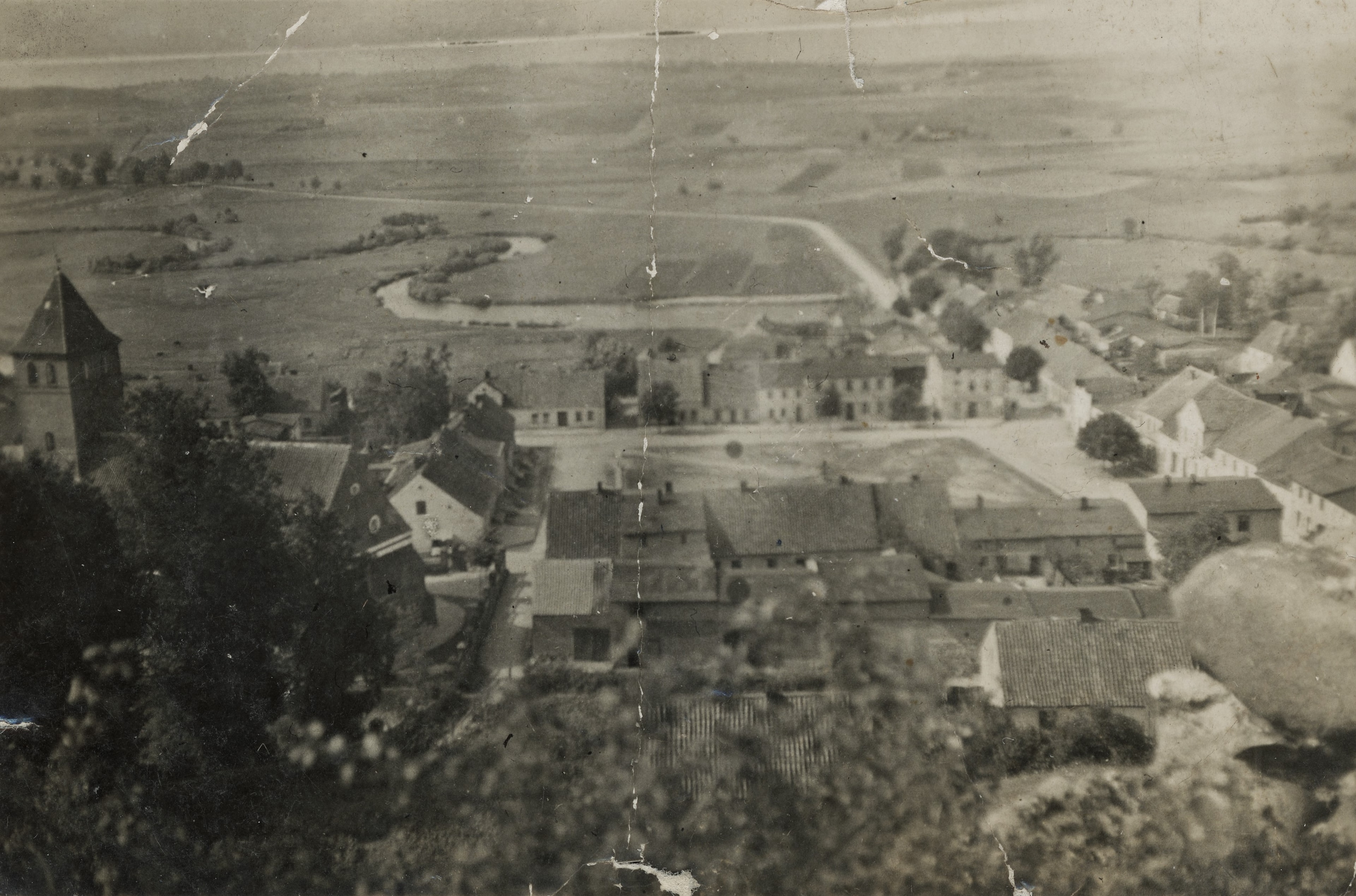Widok z lotu ptaka na historyczne miasto z wieżą kościelną, otaczającymi budynkami i otwartymi polami w tle. Obraz wydaje się stary i lekko wyblakły.