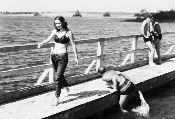Kobieta w dżinsach i staniku bikini spaceruje po drewnianym pomoście nad wodą, podczas gdy dwóch mężczyzn wychodzi z wody za nią.