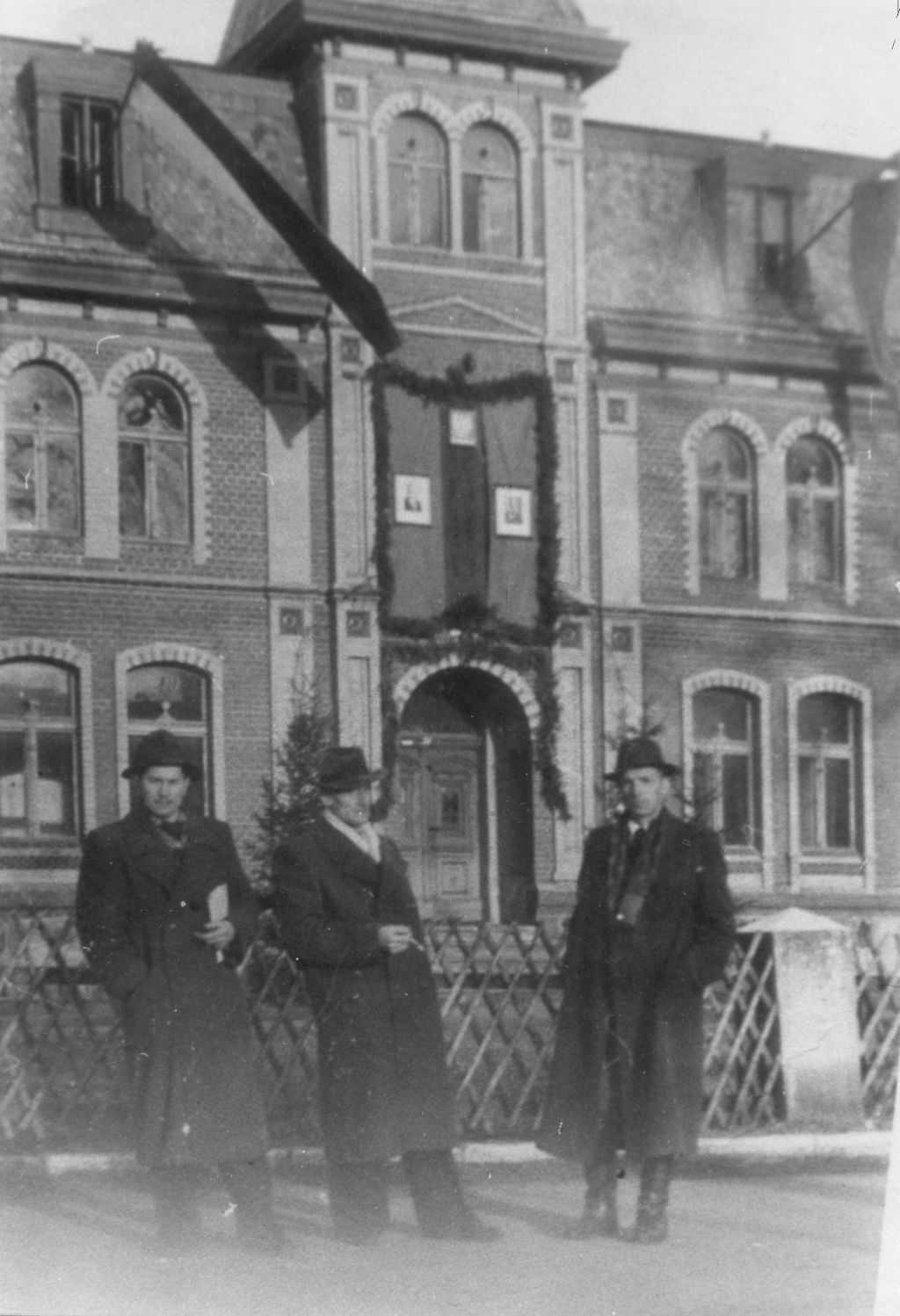 Trzech mężczyzn w płaszczach i kapeluszach stoi przed ceglanym budynkiem z ozdobnymi oknami i udekorowanym wejściem.
