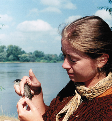 Dziewczyna trzyma w dłoniach małego ptaszka - kapturkę. Ptaszek ma szare upierzenie i czarną czapeczkę. Jest lato, w tle rozciąga się jezioro. Link do podstrony z tym zdjęciem