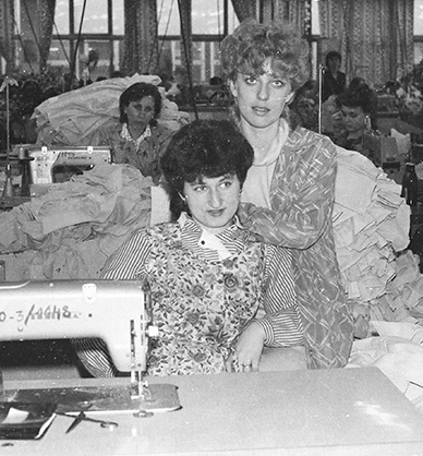 Czarno-białe zdjęcie. Wnętrze zakładu odzieżowego. Na pierwszym planie pozują dwie kobiety. Jedna siedzi przy stole, druga stoi za nią i opiera się o ramiona siedzącej. Na stole stoi maszyna do szycia. W tle widać pracujące kobiety, stosy ubrań i maszyny do szycia. Link do wyników wyszukiwania hasła zakład odzieżowy