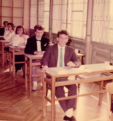 Zdjęcie kolorowe. Przedstawia uczniów podczas egzaminu maturalnego. Po prawej stronie widać duże okna. W tle drabinki gimnastyczne. Na zdjęciu widać pionowy rząd ławek. Na pierwszym planie dziewczyna w białej bluzce.Pod szyją ma czarną kokardę. Dziewczyna ma długie włosy. Patrzy w ławkę. Za nią siedzi chłopak w garniturze. Ma niebieski krawat. Patrzy w obiektyw. Uśmiecha się. W tle widać innych uczniów. Link do wyników wyszukiwania hasła matura