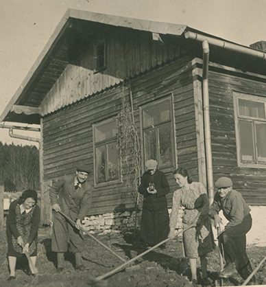 Cztery osoby przekopują ziemię w ogródku. Pomiędzy nimi ksiądz robi zdjęcie. Za nimi stoi drewniany dom. Ściany domu porasta winorośl pozbawiona liści. Link do podstrony z tym zdjęciem