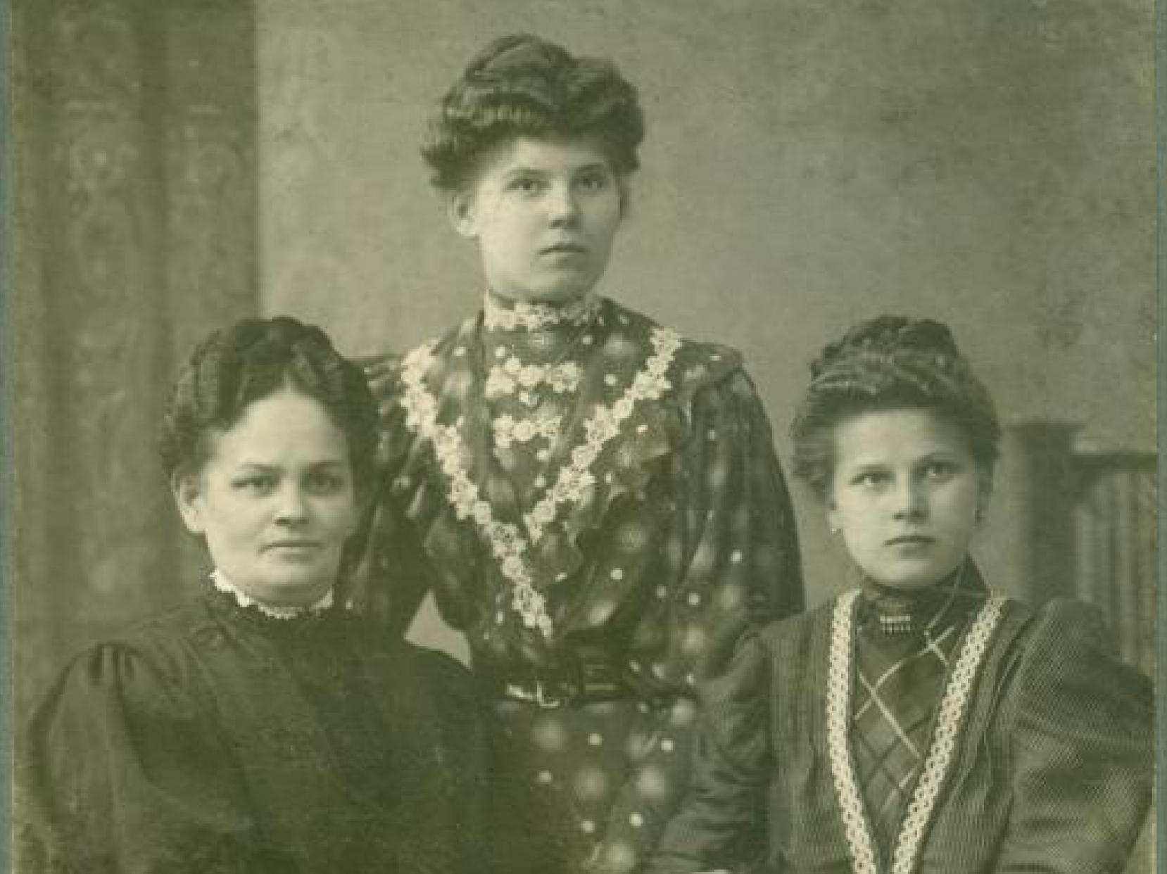 Portret trzech kobiet z początku XX wieku, ubranych w eleganckie długie suknie. Wszystkie kobiety mają upięte włosy. Dwie siedzą przy stole, jedna stoi pomiędzy nimi. Na stole leży obrus i stare pisma. Zdjęcie wykonane w atelier fotograficznym. Link do strony z artykułem zainspirowanym Zbiorami Społecznymi