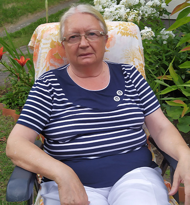 Portret starszej kobiety. Siedzi na krześle ogrodowym wśród kwiatów, patrzy prosto na widza. Link do podstrony z nagraniem audio