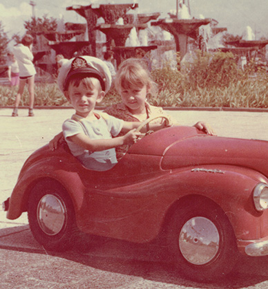 Chłopiec i dziewczynka pozują do zdjęcia w czerwonym samochodziku. Chłopiec trzyma kierownicę, na głowie ma kapitańską czapkę. Tablica rejestracyjna auta ma numer 1983. Jest lato, w tle tryska wodą fontanna. Link do podstrony z tym zdjęciem