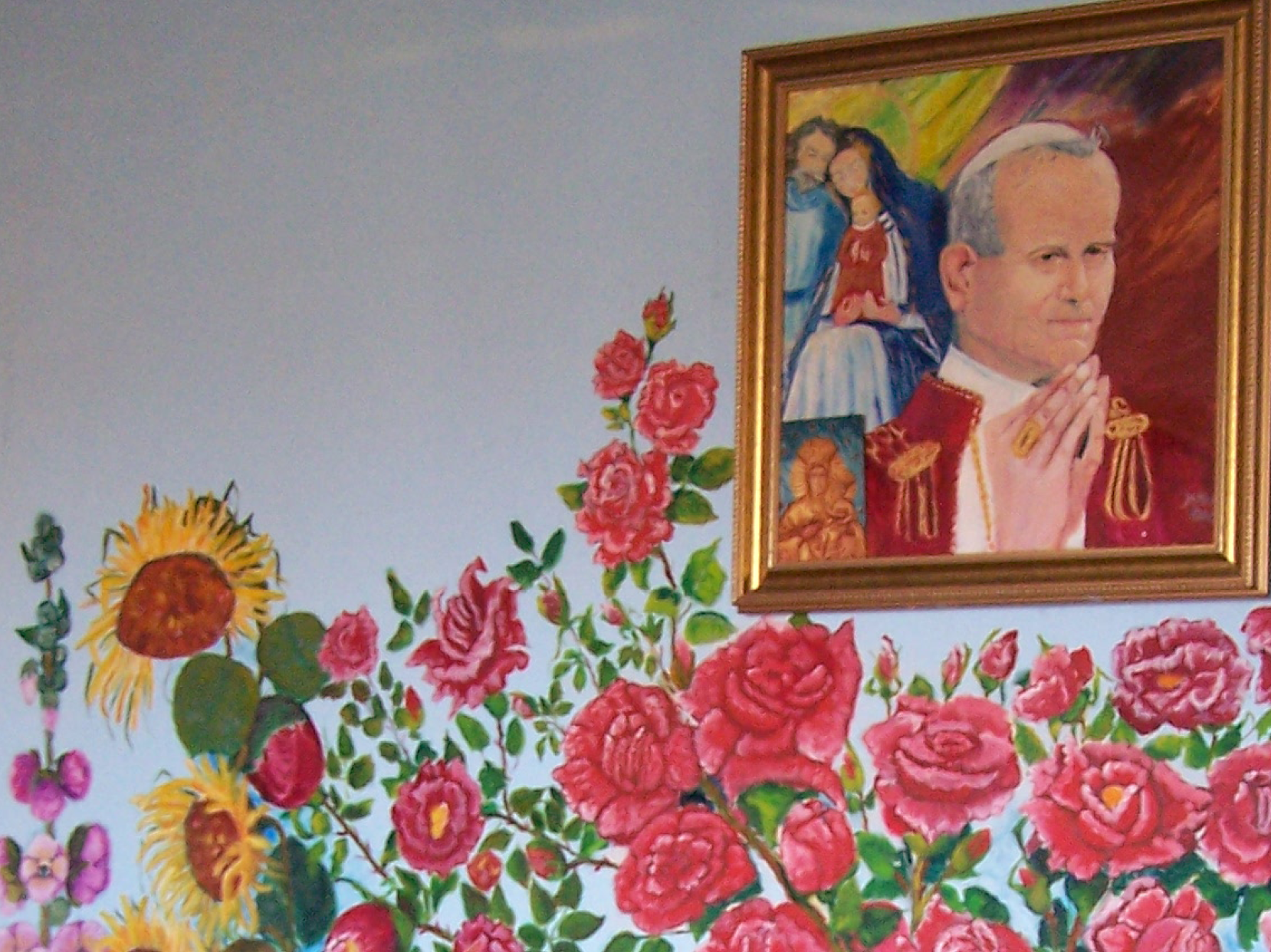 Mural na ścianie domu. W centralnej części, pomiędzy kwiatami wisi obraz przedstawiający portret papieża Jana Pawła II, z rękami złożonymi do modlitwy. Po lewej stronie Józef, Maria i Jezusek. W dolnym prawym rogu motyw Matki Boskiej Częstochowskiej. Link do strony z artykułem zainspirowanym Zbiorami Społecznymi
