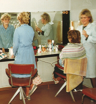 Wnętrze salonu fryzjerskiego. Uśmiechnięte fryzjerki pozują do zdjęcia. Dwie klientki siedzą przed lustrem na obrotowych fotelach. Blat stołu jest zastawiony kosmetykami i przyborami fryzjerskimi. Link do podstrony z tym zdjęciem