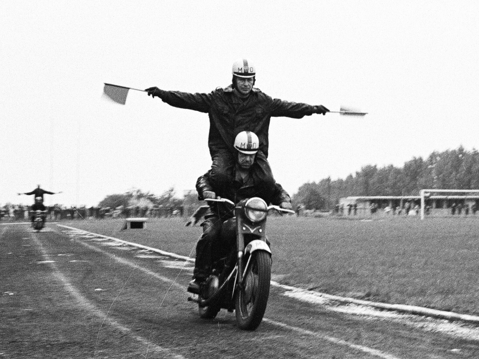 Na pierwszym planie motocyklista jadący w sposób popisowy. Przyjmuje pozycję jaskółki. Obiema dłońmi trzyma kierownicę, a tylko jedną nogą dotyka siedzenia, drugą trzyma w powietrzu. W tle widzowie obserwujący popis. Link do strony z artykułem zainspirowanym Zbiorami Społecznymi