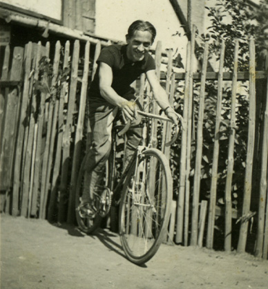 Czarno-białe zdjęcie przedstawiające mężczyznę. Jest ubrany w ciemną bluzkę z krótkim rękawem i jasne spodnie. Jedzie na rowerze. Jest pochylony lekko do przodu. Uśmiecha się. Za mężczyzną widać wysoki, drewniany płot. Za płotem znajdują się drzewa i budynki. Link do wyników wyszukiwania hasła rower