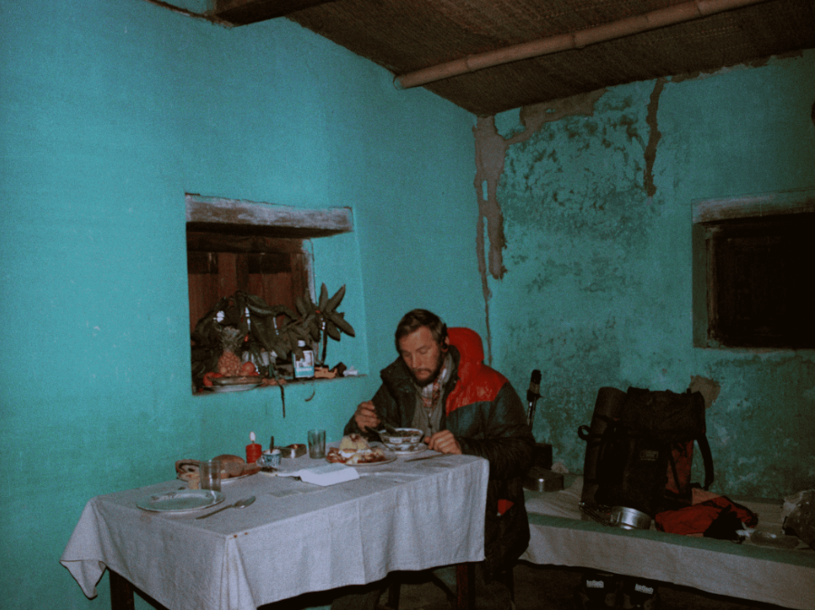 Osoba w zimowej kurtce siedzi przy stole z talerzami jedzenia w rustykalnym pokoju z turkusowymi ścianami i małymi oknami. Duży plecak leży na podłodze niedaleko.