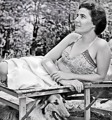 Czarno-białe zdjęcie przedstawiające kobietę. Kobieta leży na leżaku. Jest ubrana we wzorzysty stanik i spódnicę. Opiera się na łokciu. Patrzy w bok. Pod leżakiem leży pies. Ma długą sierść i otwarty pysk. W tle widać drzewa oraz krzewy. Link do wyników wyszukiwania hasła mama